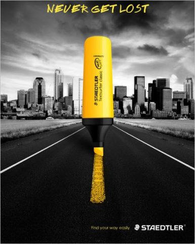 Resaltador amarillo de marca STAEDTLER traza una linea en medio de una carretera. En el fondo se ve una ciudad. Se hace énfasis en el producto siendo lo único que tiene color.
